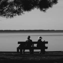 A couple sitting apart from each other on a bench beside a lake.