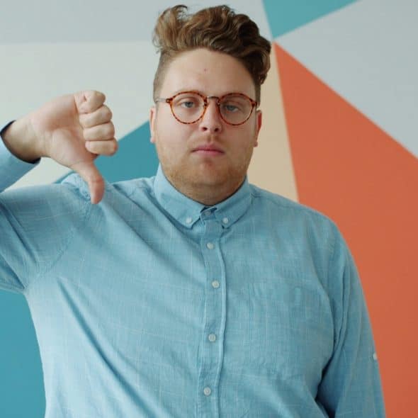A man with glasses gives a thumbs-down gesture in front of a geometric colorful wall.