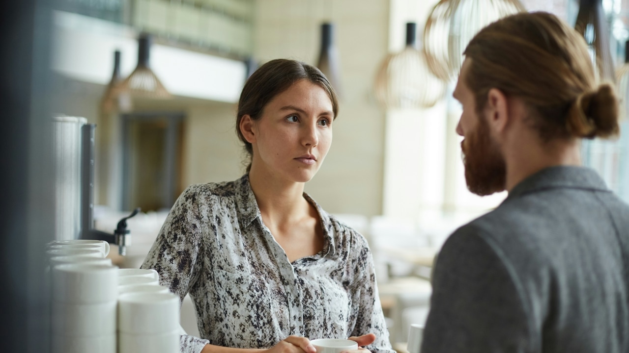 A woman in a patterned blouse looks at a man with a beard and a hair bun.