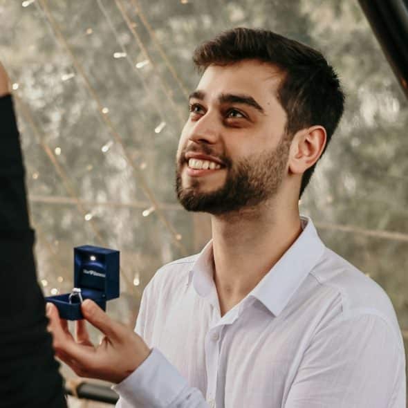 A man kneels and holds an open blue ring box toward a woman in black.