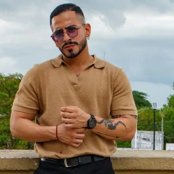 A man with tattoos and sunglasses wearing a tan polo shirt stands against a city backdrop.