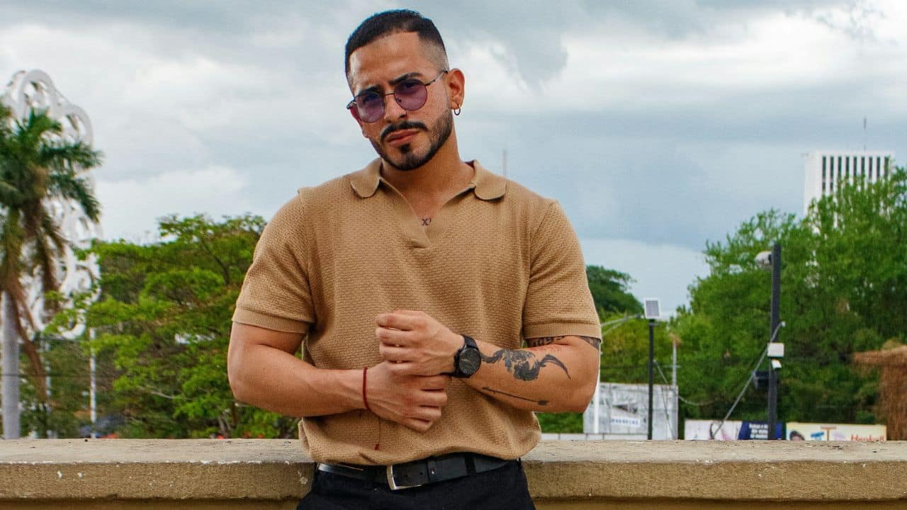 A man with tattoos and sunglasses wearing a tan polo shirt stands against a city backdrop.
