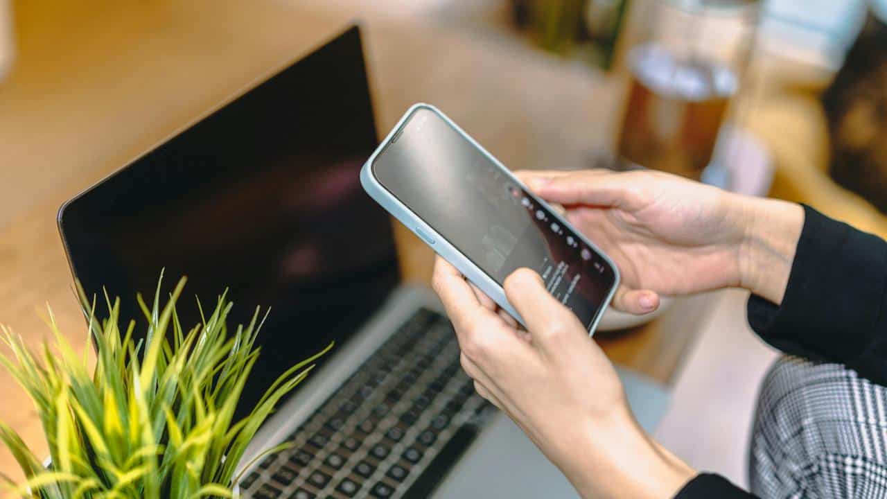 A person holding a smartphone next to a laptop and a small plant on a desk.