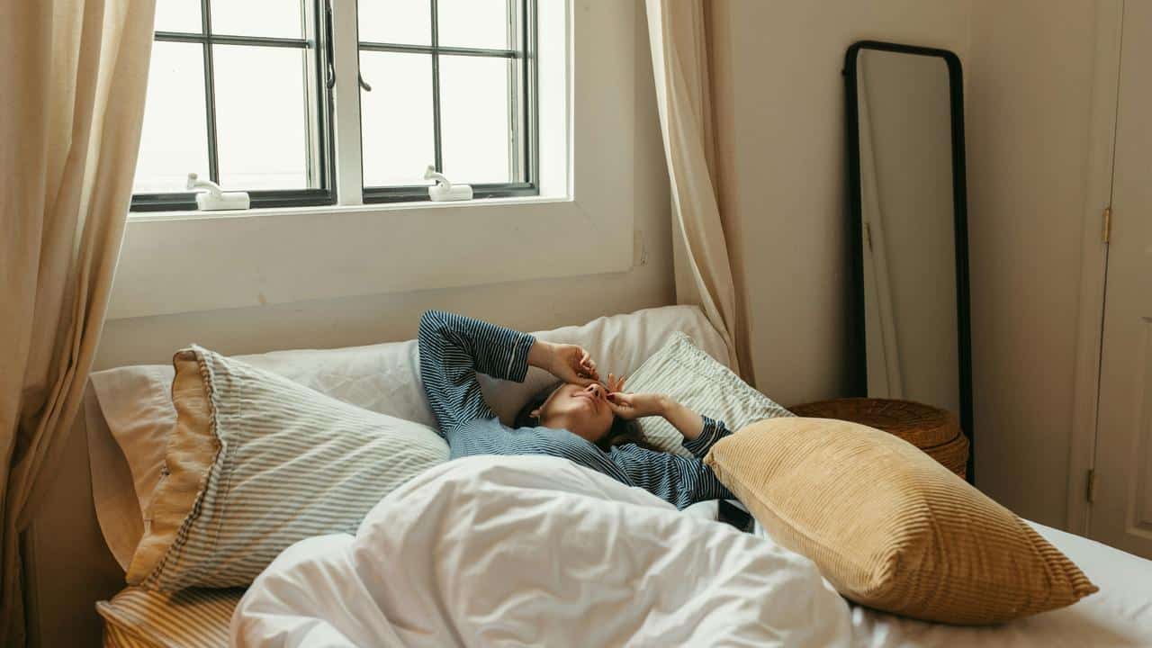 A person lying in bed with pillows, covering their eyes near a window.
