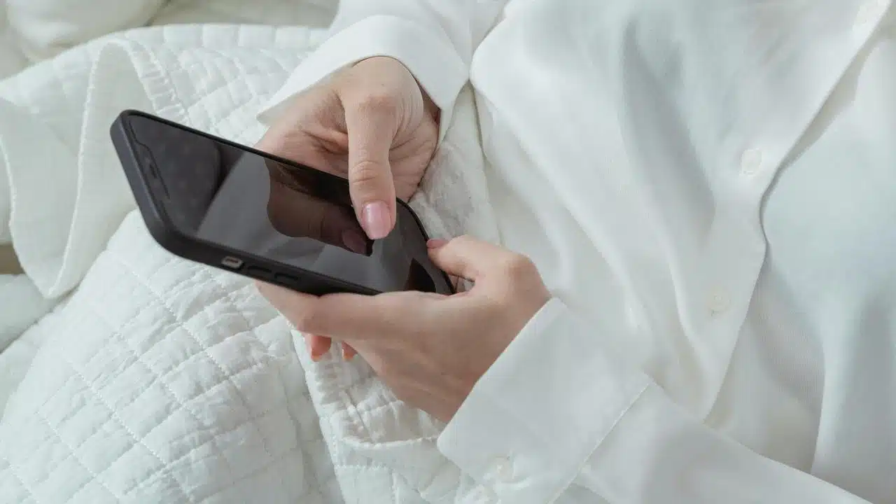 A person in a white shirt holding a smartphone while sitting on a bed.