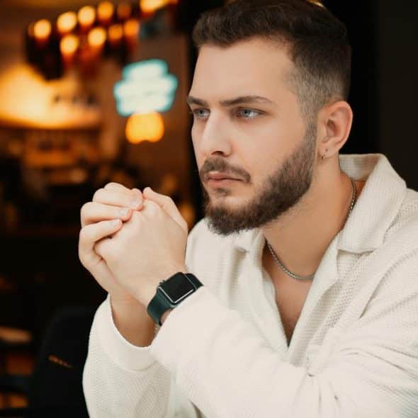 A bearded man in a light sweater sitting thoughtfully at a table