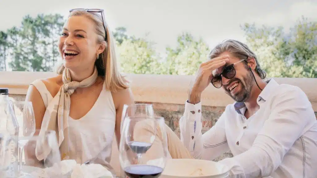 A smiling woman and a laughing man sit at a table with wine glasses outdoors.