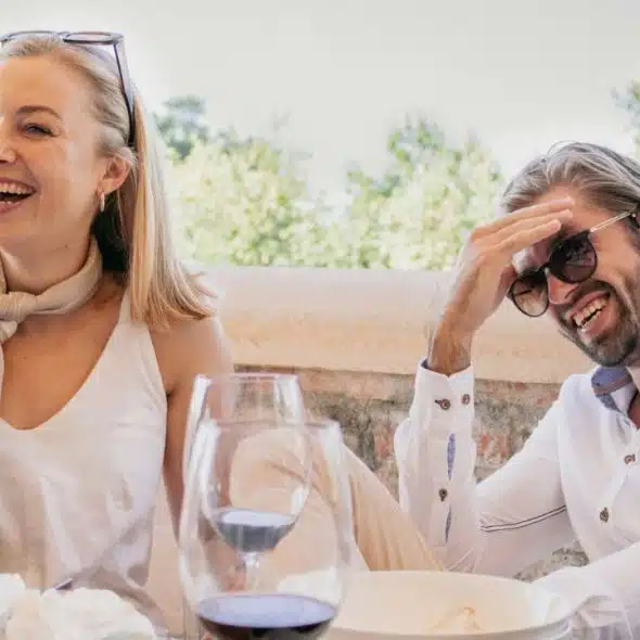 A smiling woman and a laughing man sit at a table with wine glasses outdoors.