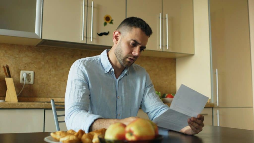 A man in a light blue shirt sits in a kitchen reading a white paper document.