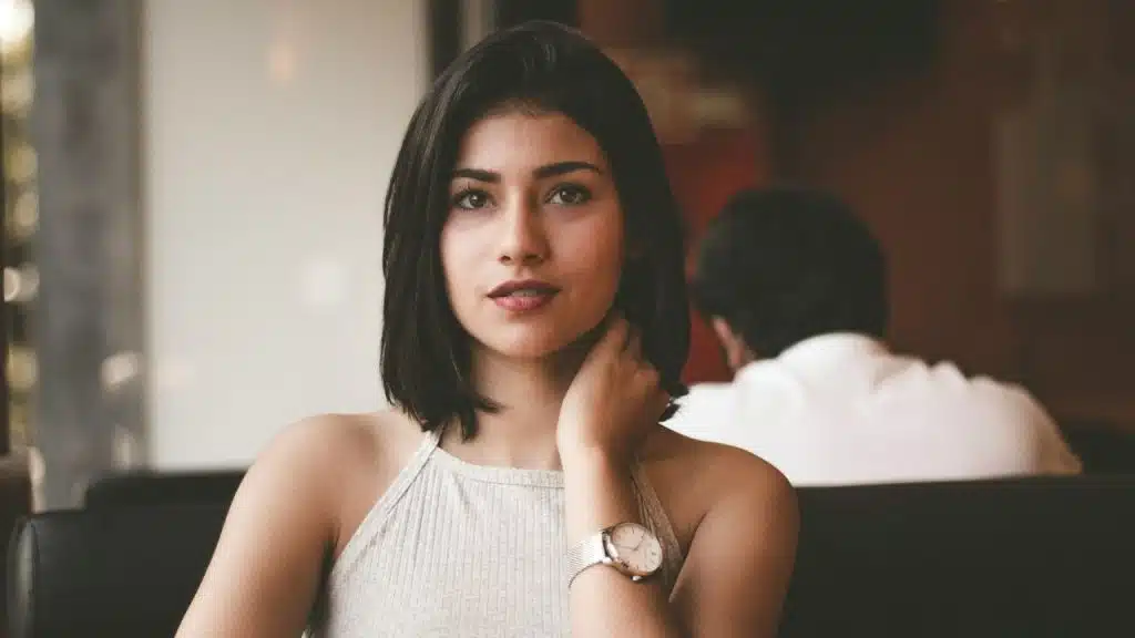 A woman with dark hair and a wristwatch looking forward while touching her neck.