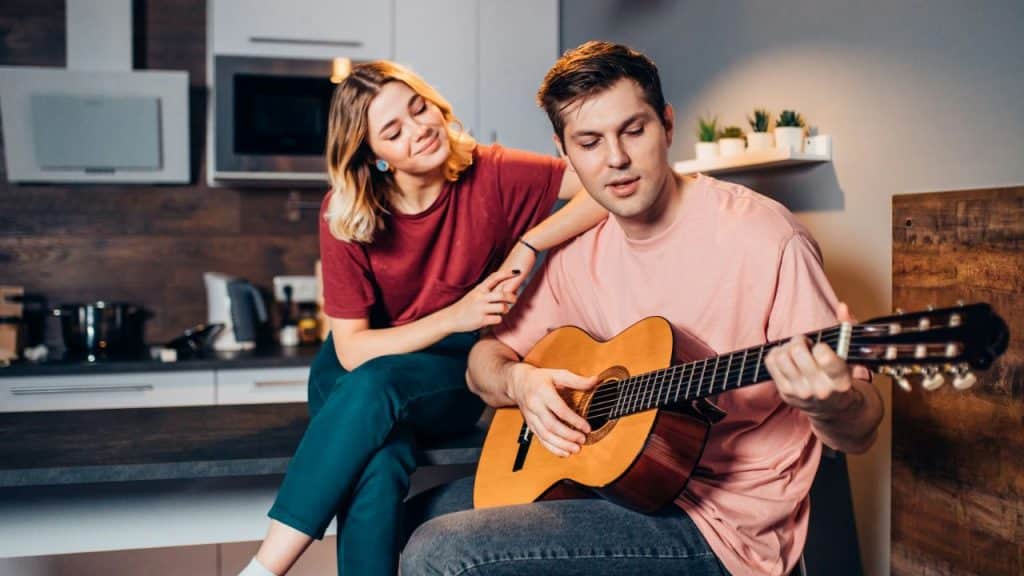 A man plays an acoustic guitar in a kitchen while a woman watches him intently.