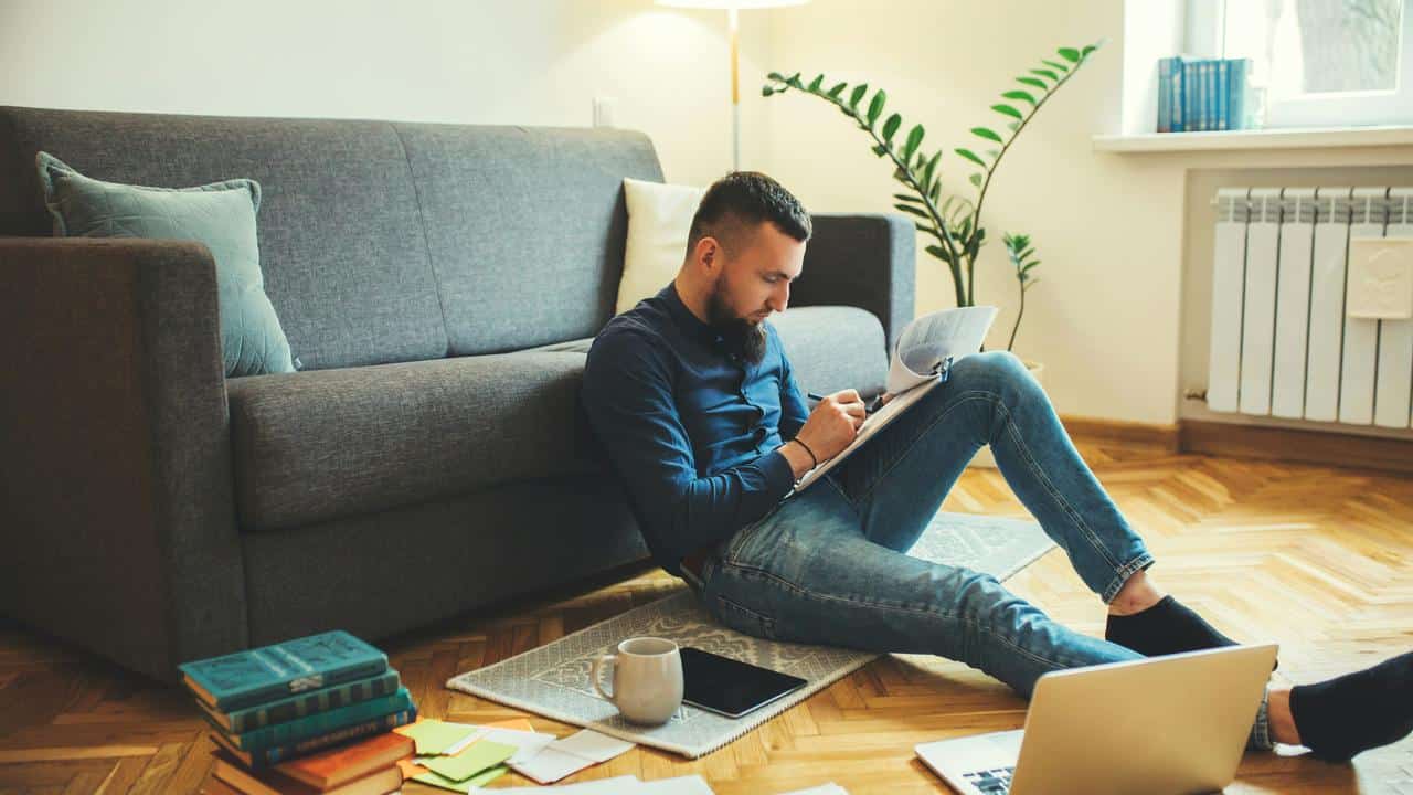 A man sitting on the floor by a sofa, reading and taking notes at home.