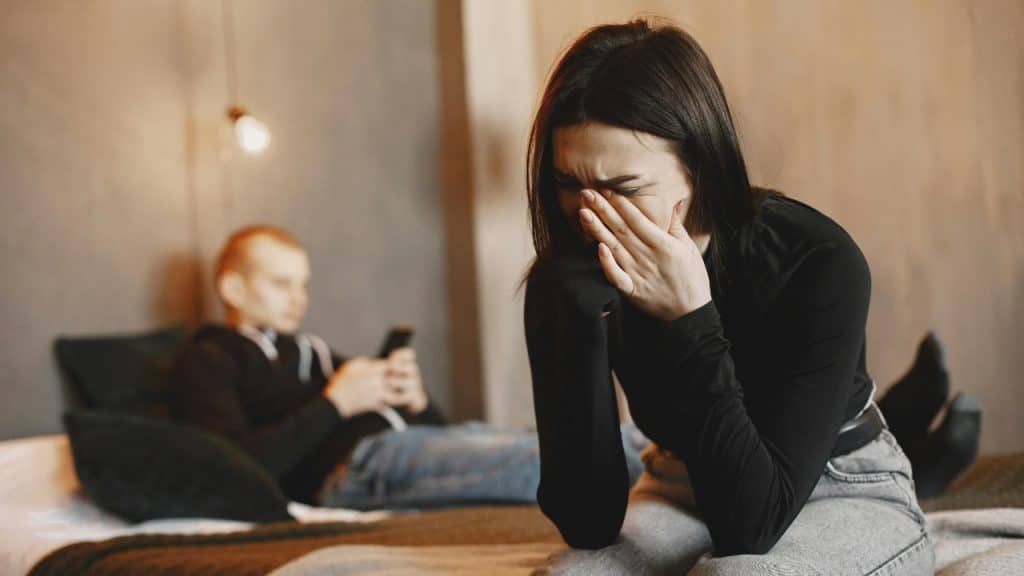 A woman sits on a bed crying while a man in the background uses his phone.