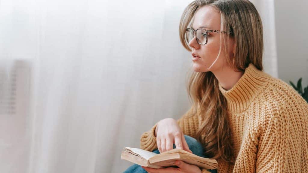 A woman wearing glasses sits indoors, holding an open book with a thoughtful expression.