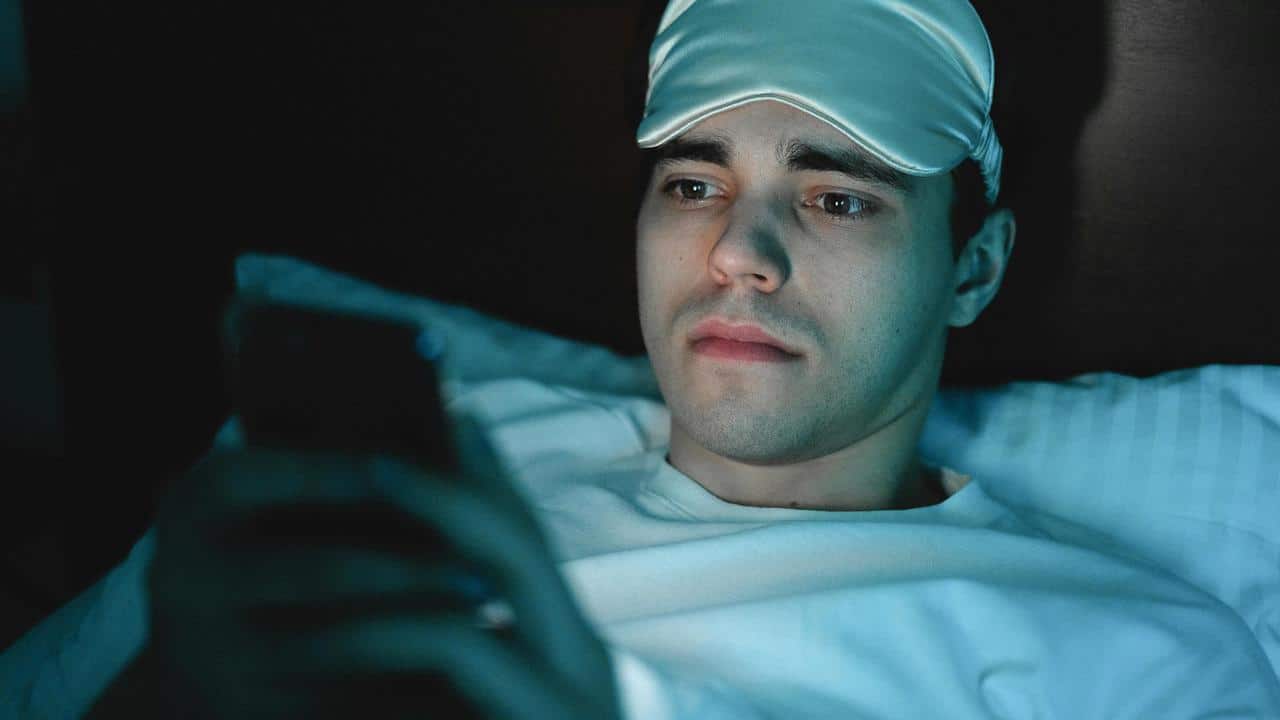 A person lying in bed wearing a sleep mask and looking at a phone.