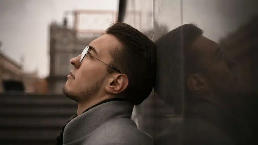 A serious man with glasses and a beard is leaning his head against a stone wall, looking up.
