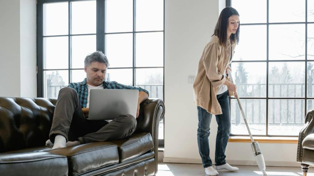 A man uses a laptop on a sofa while a woman vacuums the floor nearby.