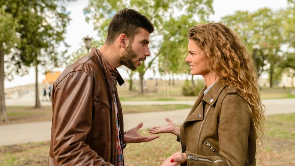 A man and woman in brown jackets stand outdoors talking with expressive hand gestures and facial expressions.