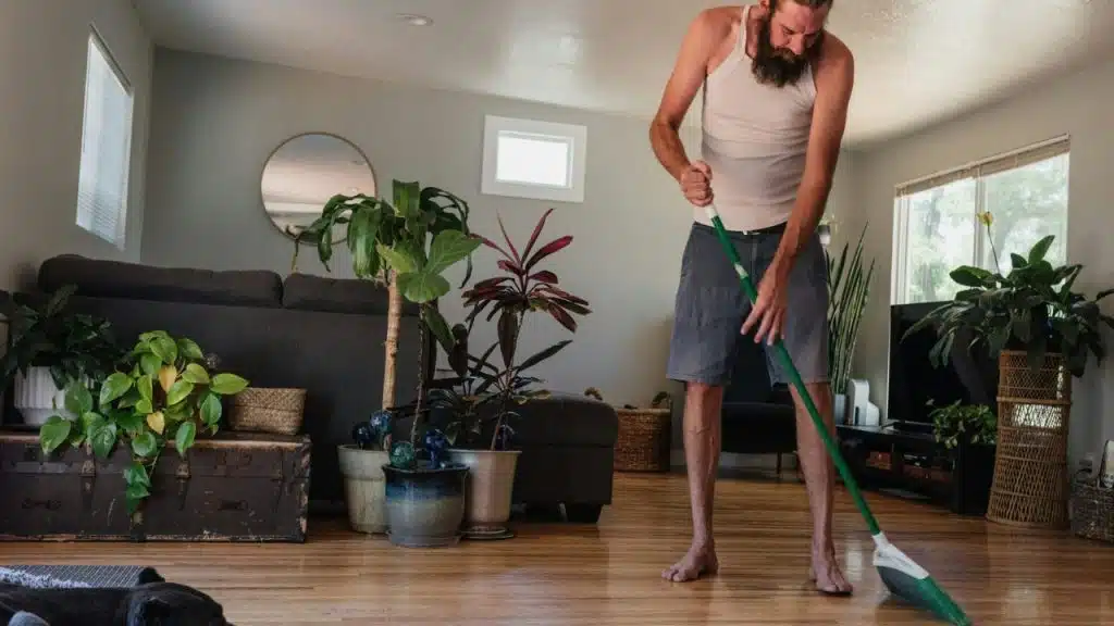 A barefoot man with a beard sweeps a wooden floor with a green broom indoors.
