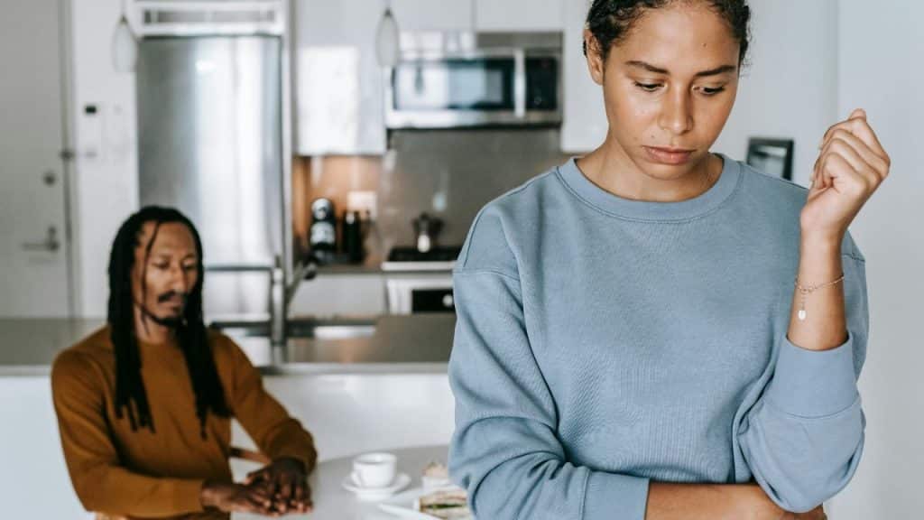 A woman looks down with arms crossed while a man sits in a kitchen background.
