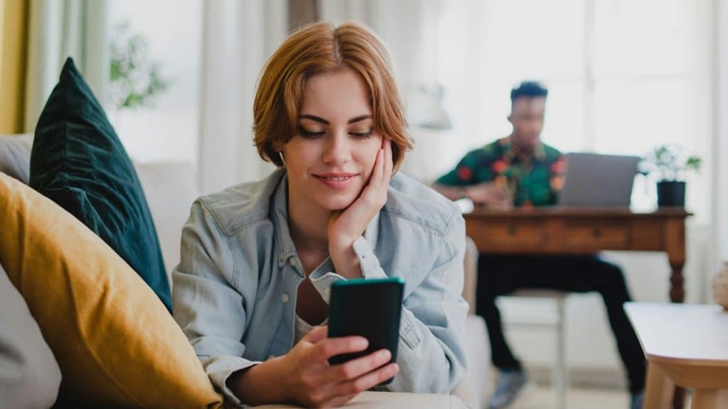 A woman smiles at a smartphone while a man works at a desk behind her.