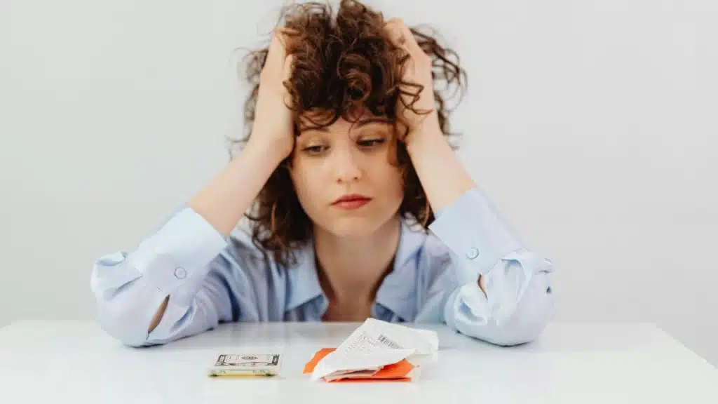 A worried woman holding her head while looking at bills and money on a table.