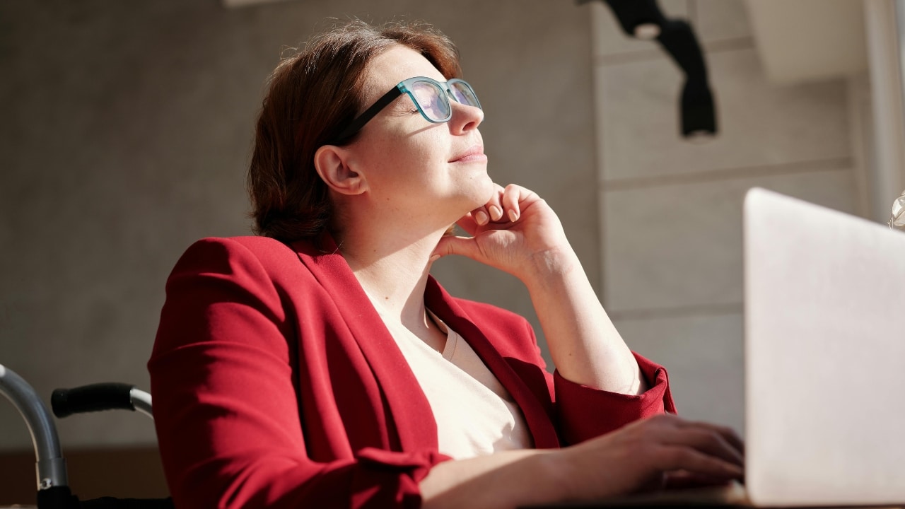 A woman in a red blazer and glasses smiles while looking up from her laptop.