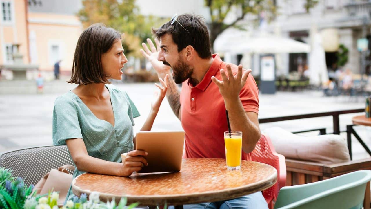A man and woman at an outdoor cafe table argue while looking at a digital tablet.