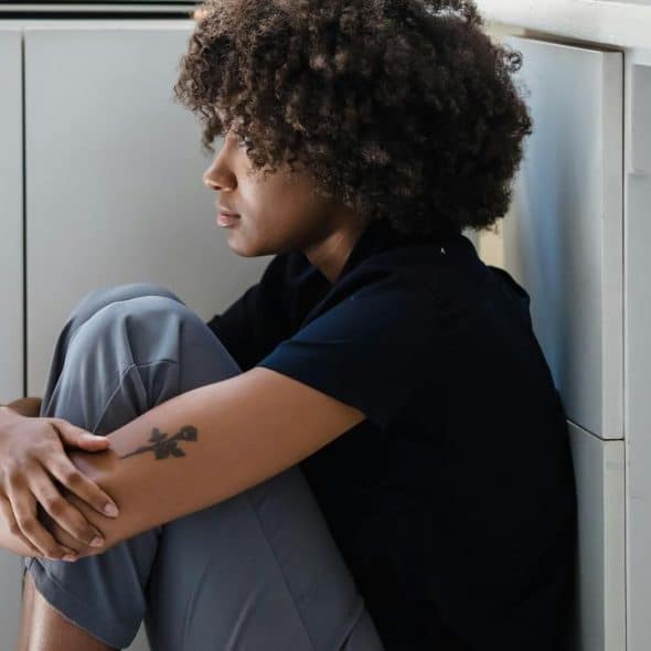 A woman sitting on the floor hugging her knees and looking away pensively.