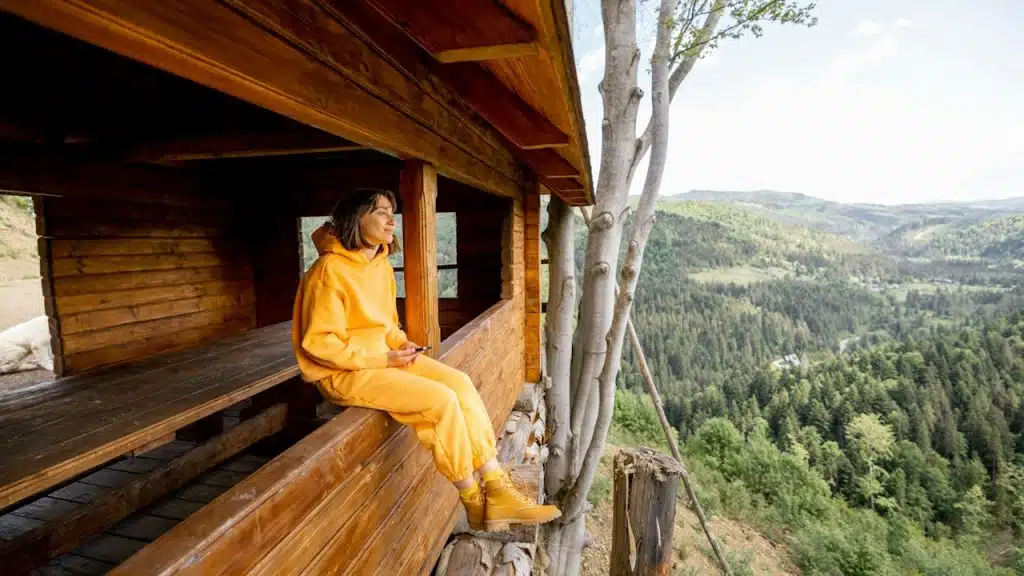 A young woman in an orange sports suit enjoys a great mountain landscape while sitting on the window of a tiny wooden tiny house.