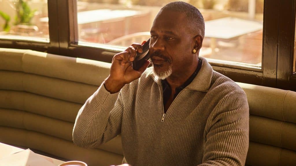 A man seated in a restaurant booth, holding a phone to his ear in warm sunlight.