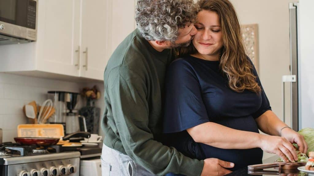 A man hugging a woman from behind as she chops vegetables in the kitchen.