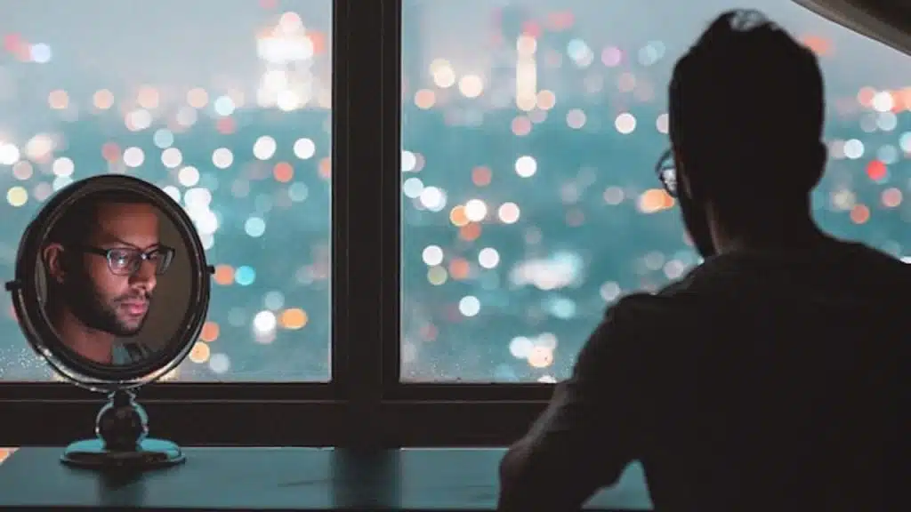 A man looks out a window at night with his face reflected in a mirror.