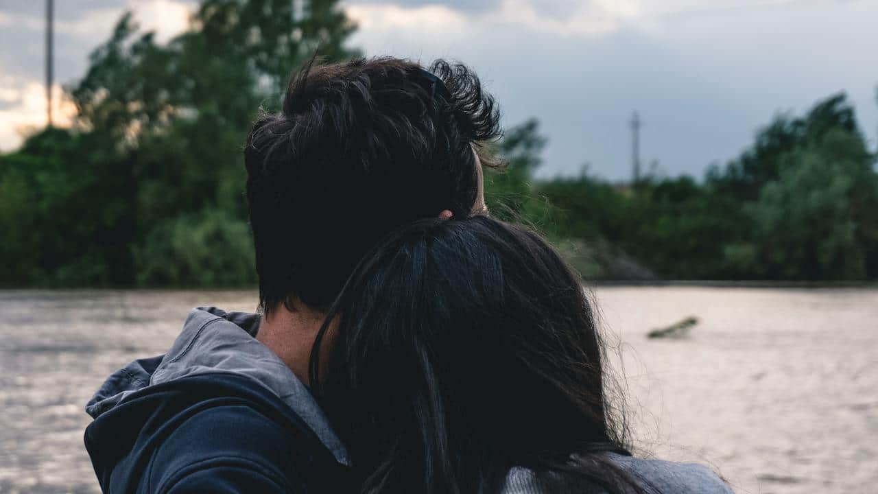 A couple embracing quietly by a river with trees in the background.