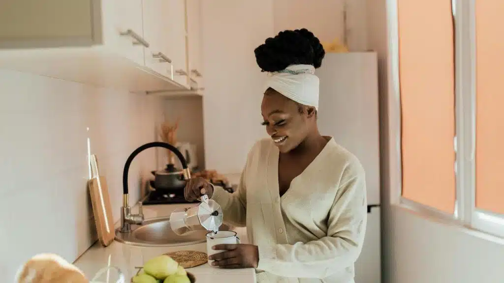 A woman stands in a bright kitchen, smiling as she pours coffee into a cup.