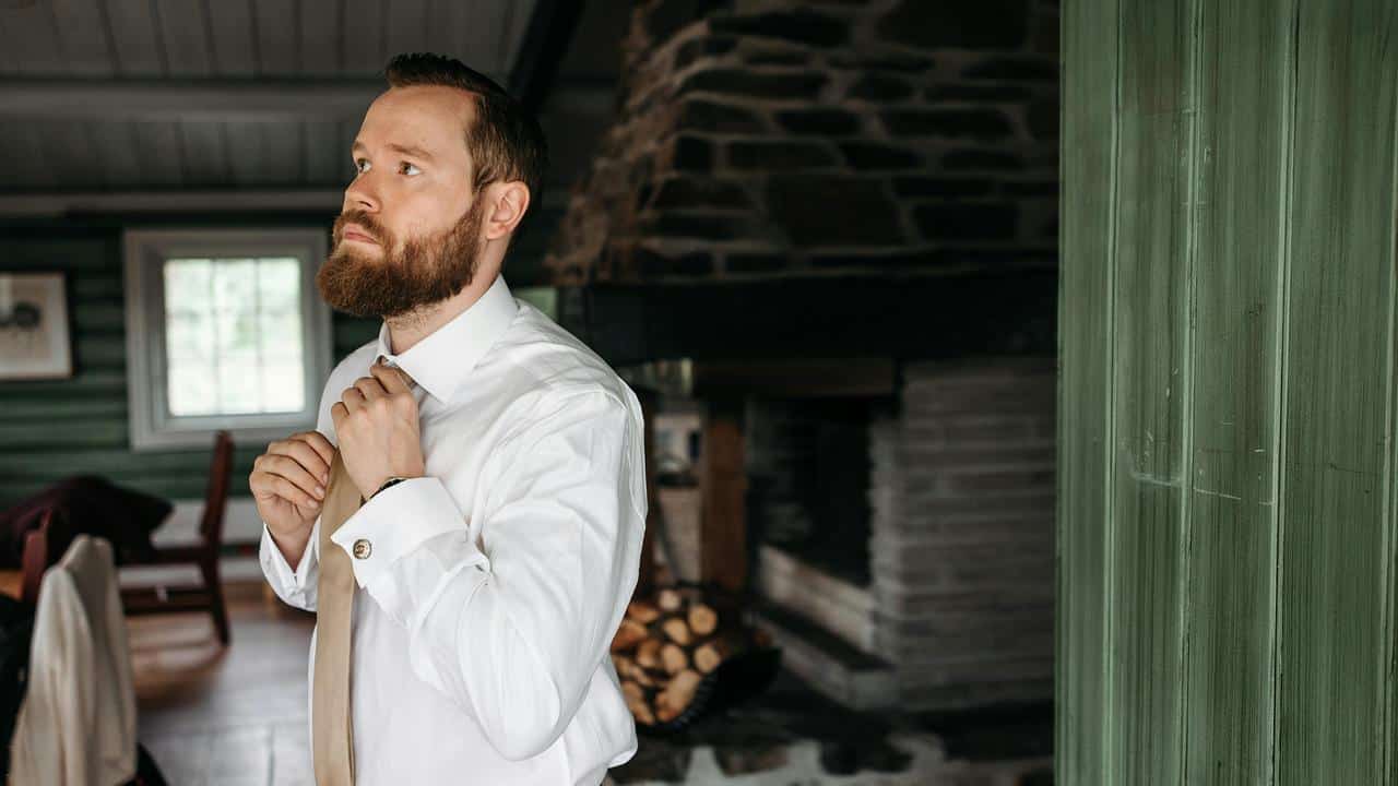 A bearded person adjusting a tie inside a rustic room.
