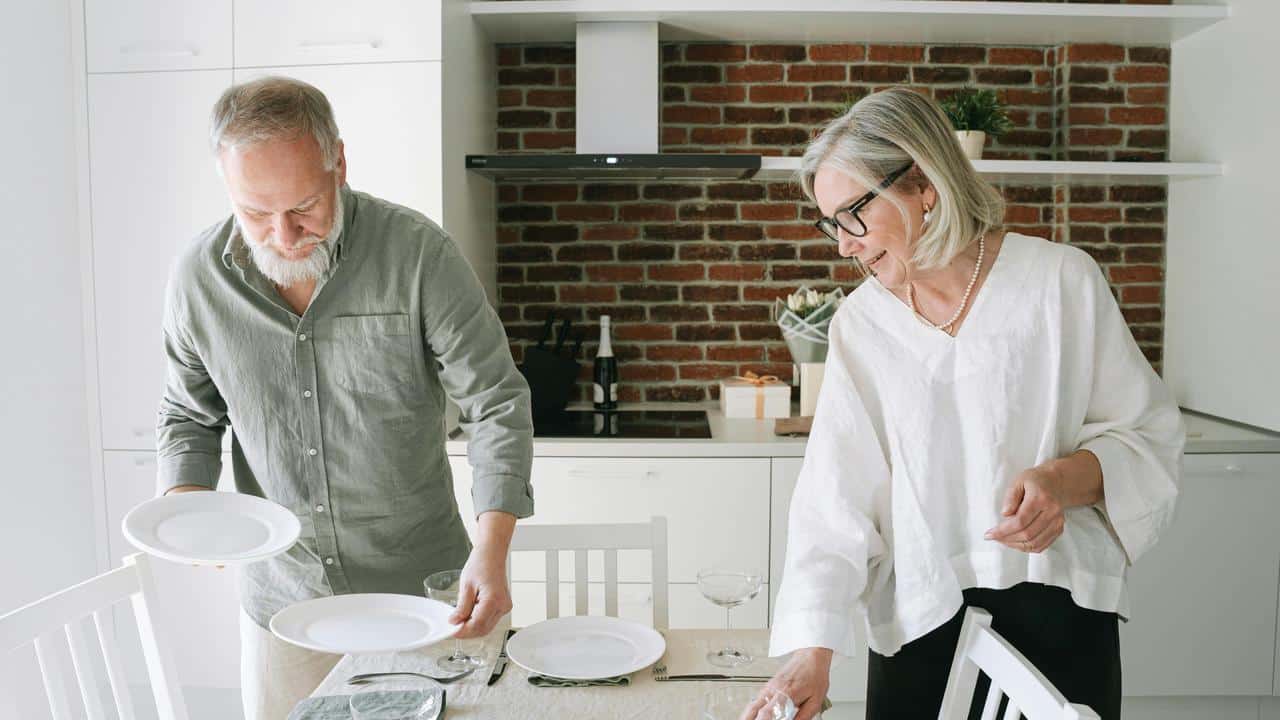 A senior couple setting the table together in a kitchen.