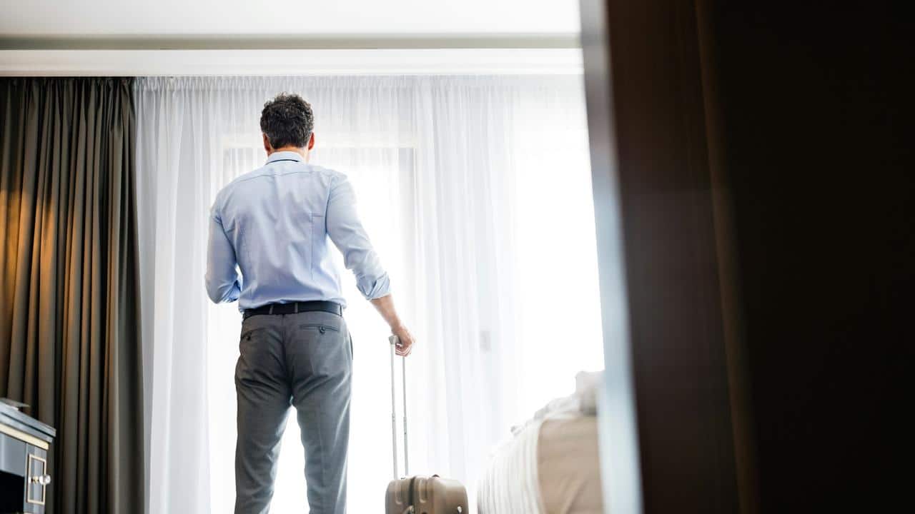 A man standing by a window with a suitcase in a hotel room.