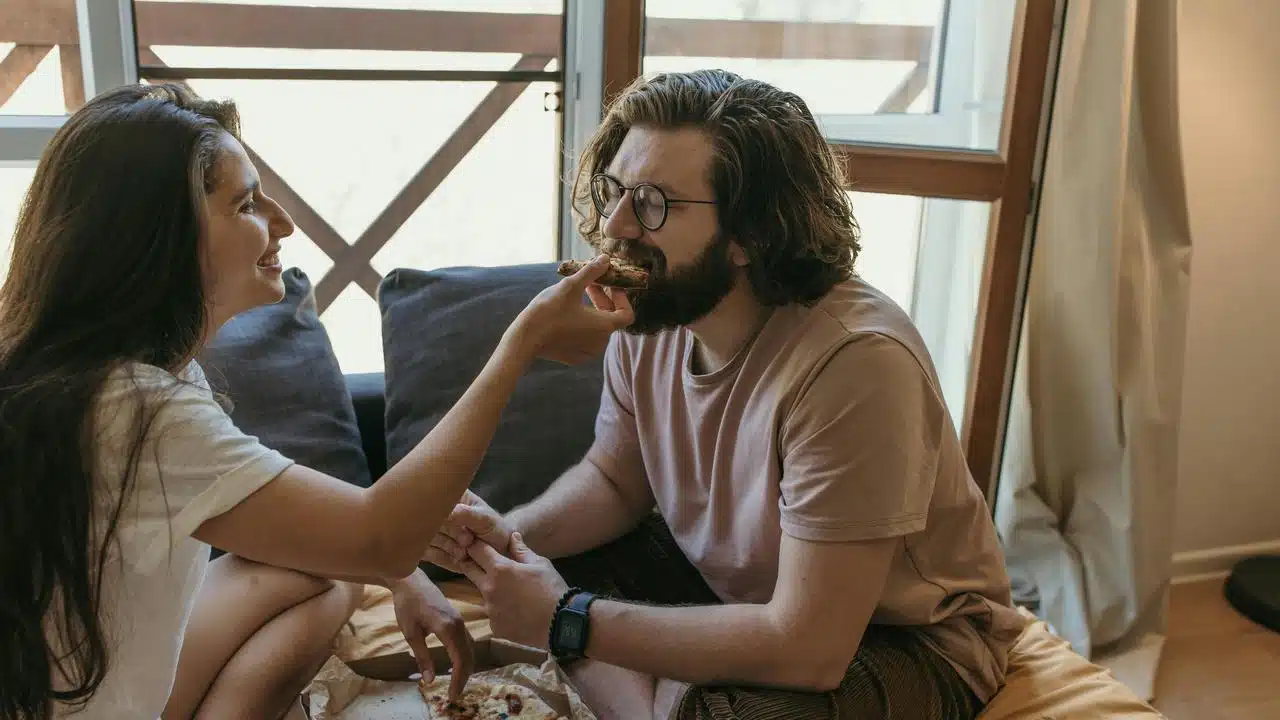 A woman feeding pizza to a man while they sit together indoors.
