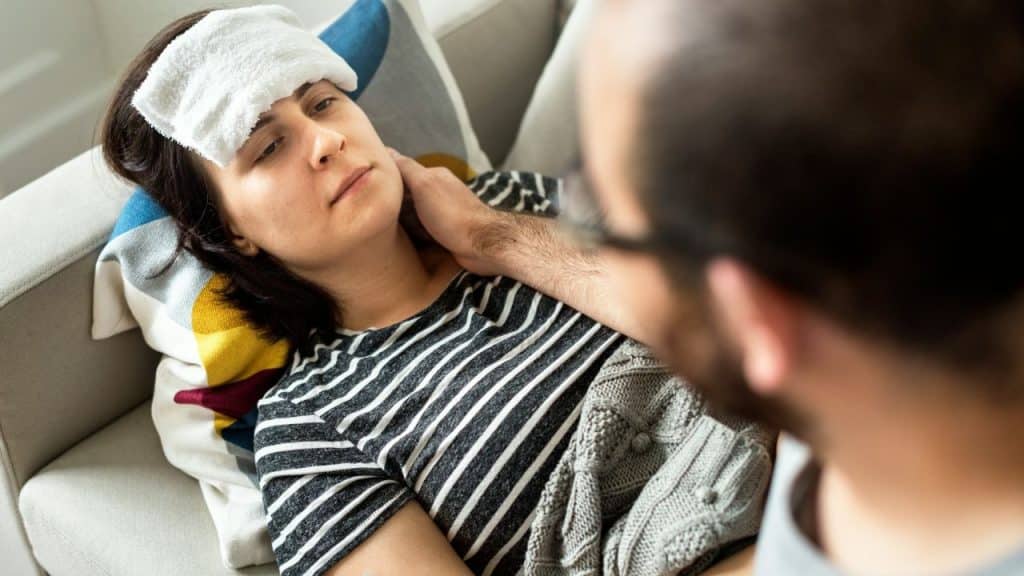 A woman lies on a sofa with a cloth on her head while a man watches.
