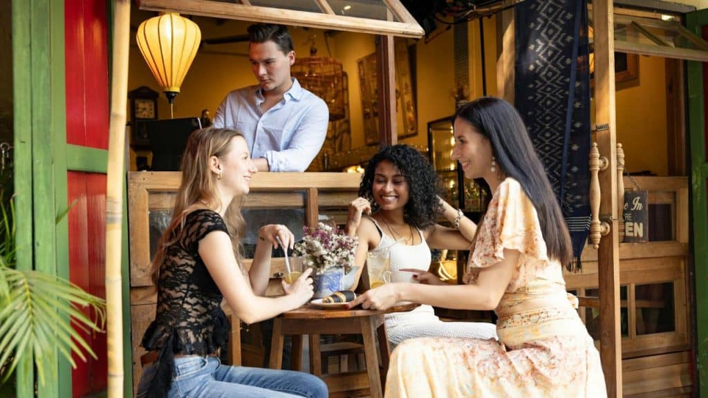 Three women sit at an outdoor table laughing while a man watches from behind a counter.