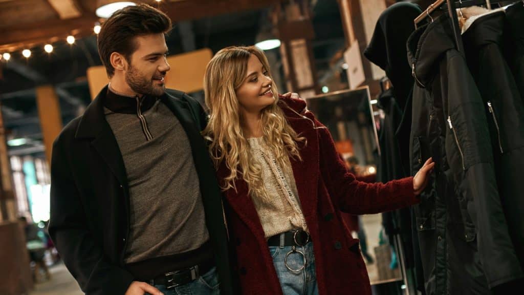A man and woman walk together while looking at dark coats hanging on a rack.