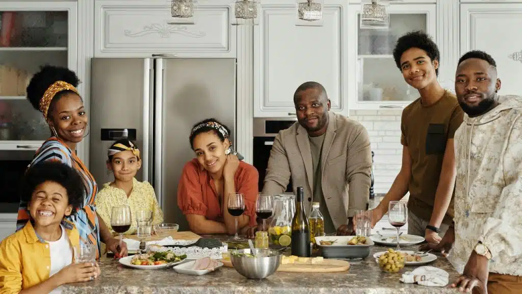 A large family group smiles together behind a kitchen island filled with various foods.