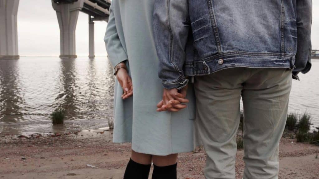 A couple holding hands by the water near a bridge.