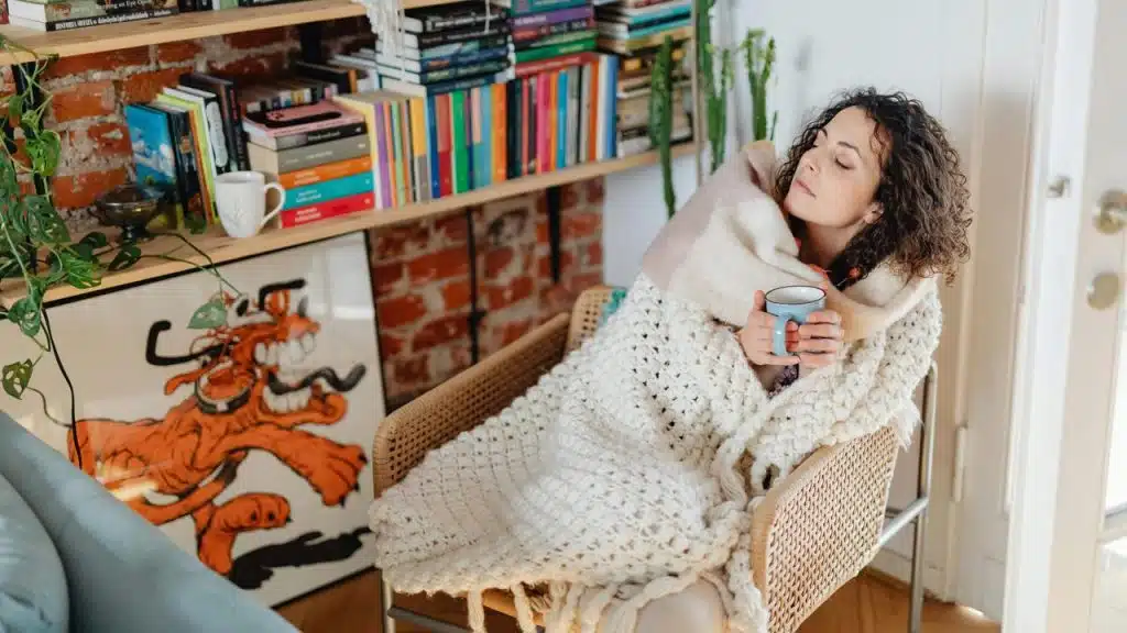 A woman wrapped in a blanket, holding a mug while relaxing in a chair beside a bookshelf.