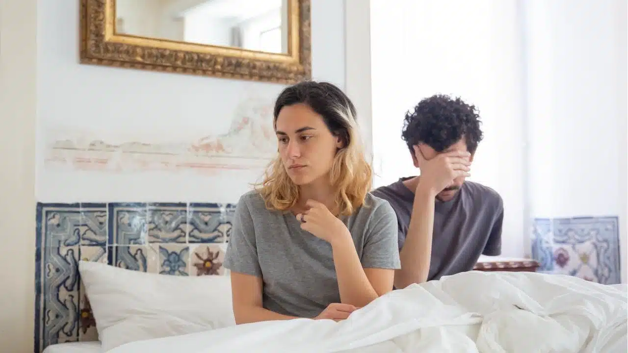 A couple sits on a bed after an argument; the woman looks sad while the man holds his hand over his face.