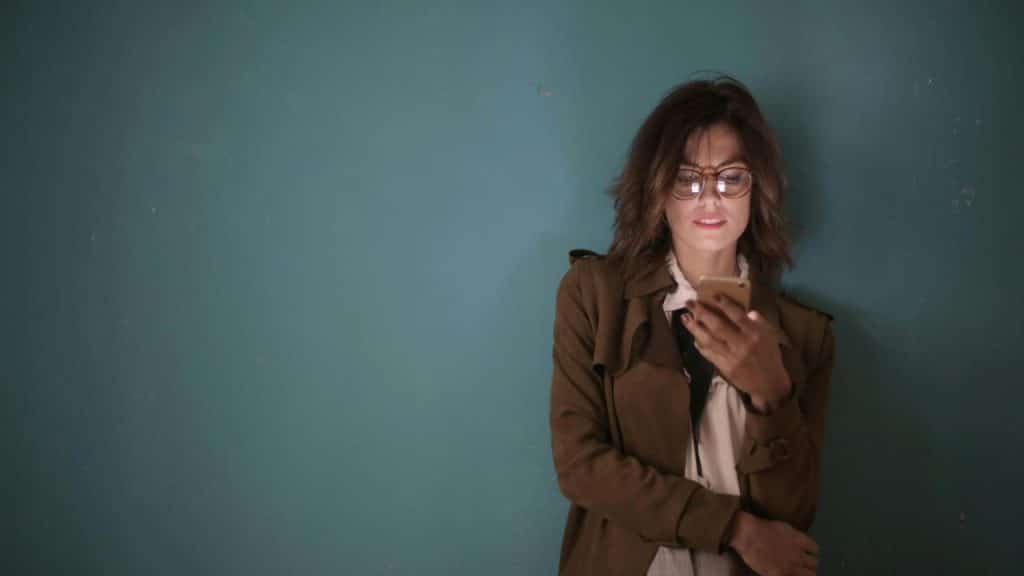A woman wearing glasses looking at her phone while standing against a blue wall.