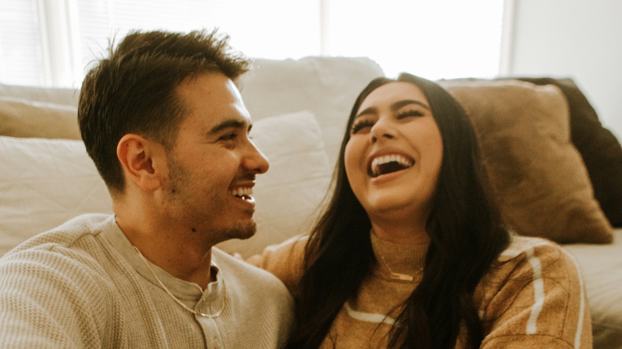 A man and woman sit closely together on a couch, laughing and looking at each other.