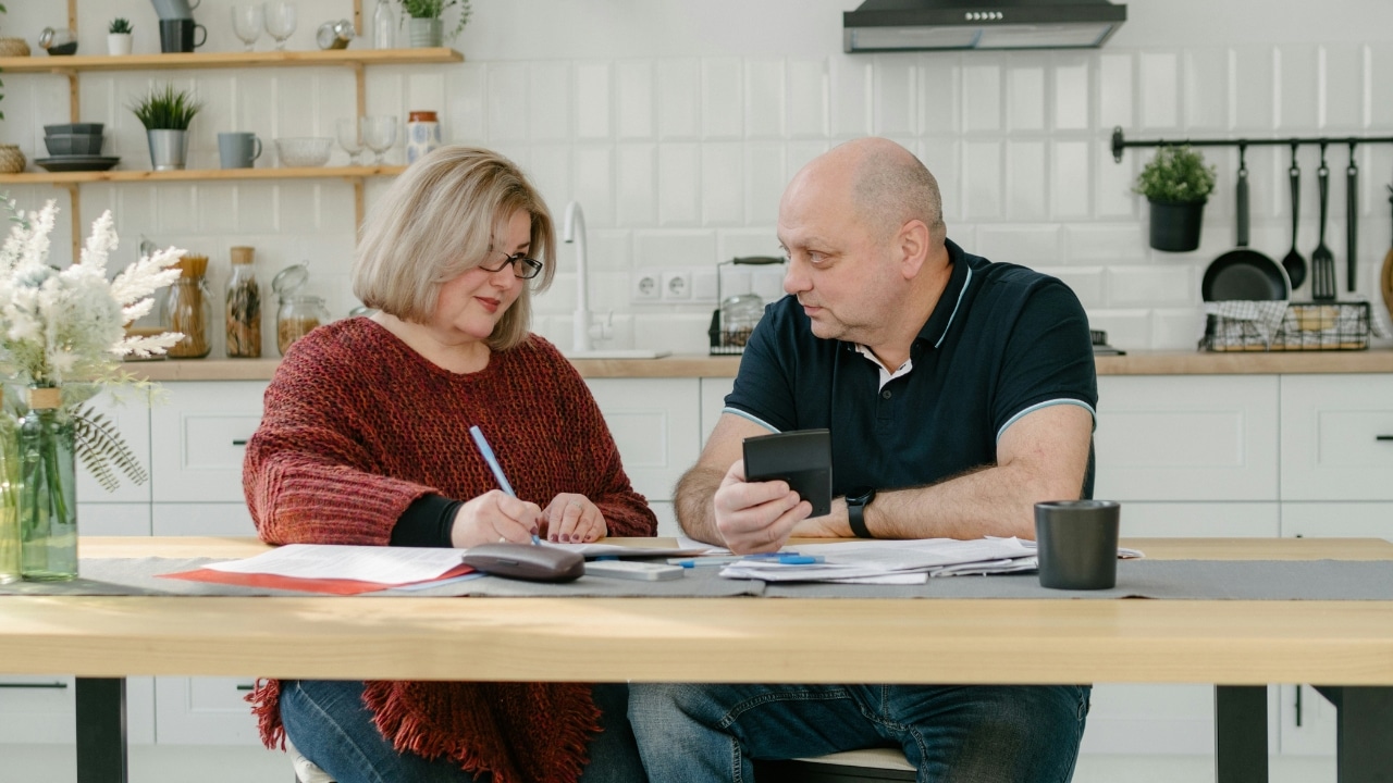 A man and woman sit at a wooden table with papers, a calculator, and a pen.