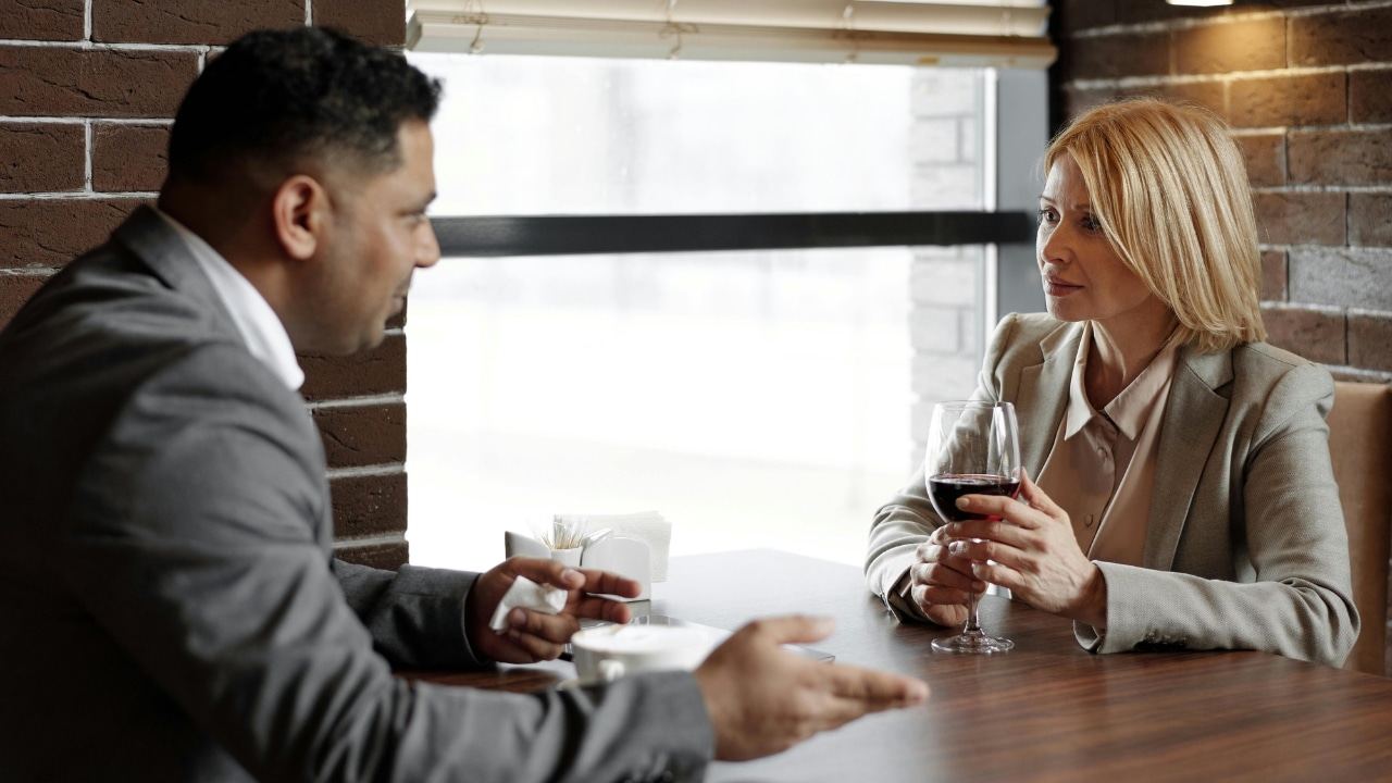 A man and woman in business attire sit at a restaurant table having a conversation.